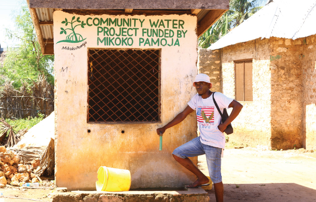 photo of Ismail Barua standing with his hand resting on a water faucet, with the words Community Water Project Funded by Mikoko Pamoja written on the water kiosk behind him
