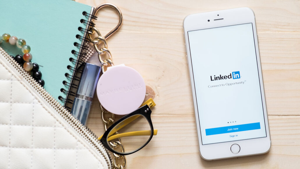 A phone on a desk showing the LinkedIn login screen, alongside a purse, notebook and other accessories