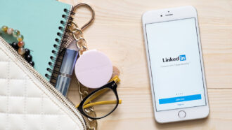 A phone on a desk showing the LinkedIn login screen, alongside a purse, notebook and other accessories