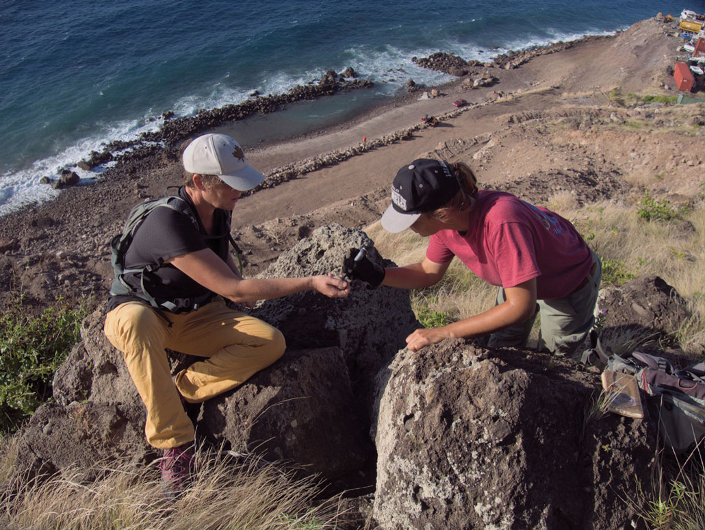Două persoane măsoară un ou de pasăre tropică cu cioc roșu pe o stâncă din Saba.
