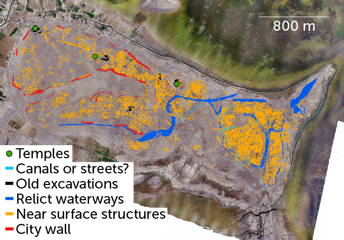 A map overlaying the ancient Mesopotamian city of Lagash on a present-day Iraq site. Three green dots indicate temples, a few light blue lines indicate possible canals or streets, several small black clusters indicate old excavations, long blue passages mostly on the right half indicate relict waterways, yellow dots heavily clustered on the right half but spread throughout the site indicate near surface structures, and red lines mostly on the left half indicate city wall