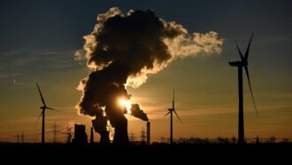 Wind turbines stand near a coal-fired power plant in Germany, with steam rising from the plant's cooling towers.
