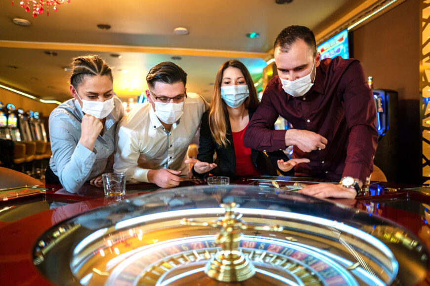 Four people with masks on, stand behind a roulette table