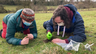 two researchers crouched on the ground in a field examining and collecting pieces of the Winchcombe meteorite