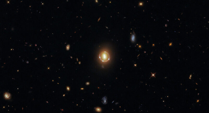 Quasar 2M1310-1714 è visibile come quattro punti di luce attorno a una luce centrale grazie alla lente gravitazionale. È un'immagine dello spazio in alto.