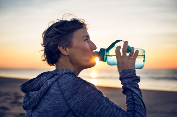 A photo of a woman drinking from a water bottle on the beach with the sun setting in the background