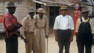 Five Black men, all wearing hats, stand near an old building