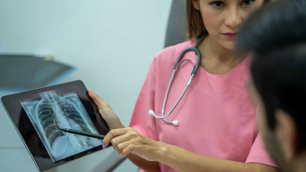 a medical professional in pink scrubs holds and points to a tablet with an x-way and talks to a patient