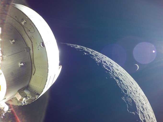 the Orion spacecraft in the foreground with the moon prominent in the background. Earth is shown as a thin crescent, as well as a large lens flare with a tiny dot in the middle, which is the sun