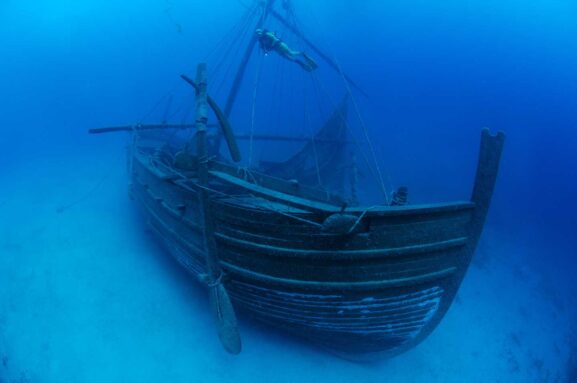 An underwater photo of a replica sunken merchant ship with a scuba diver swimming above.