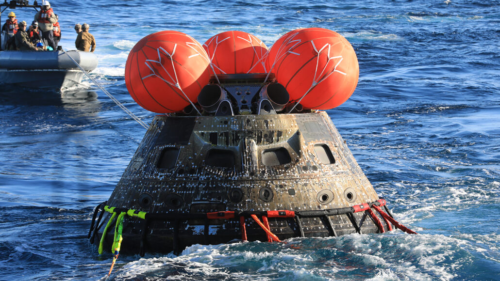 the uncrewed space capsule Orion floating in the Pacific Ocean, with round red airbags atop it as a crew in a boat in the background retrieves the capsule