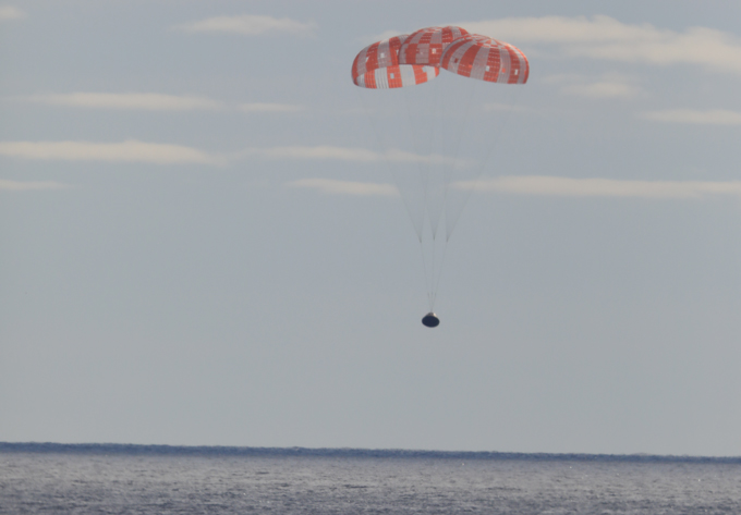 the Orion spacecraft descends to the Pacific Ocean, slowed by a large red-and-white parachute