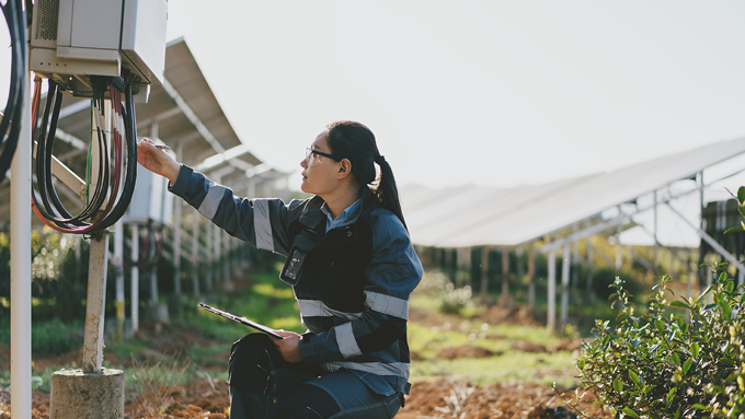 A photo of a woman kneeling next to an electrical box with solar panels in the background.