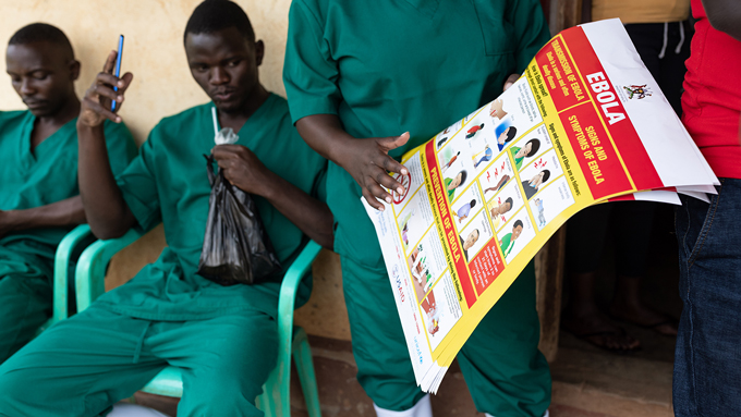 Foto de dos voluntarios de la Cruz Roja sentados y un voluntario de pie sosteniendo y señalando una infografía sobre el ébola