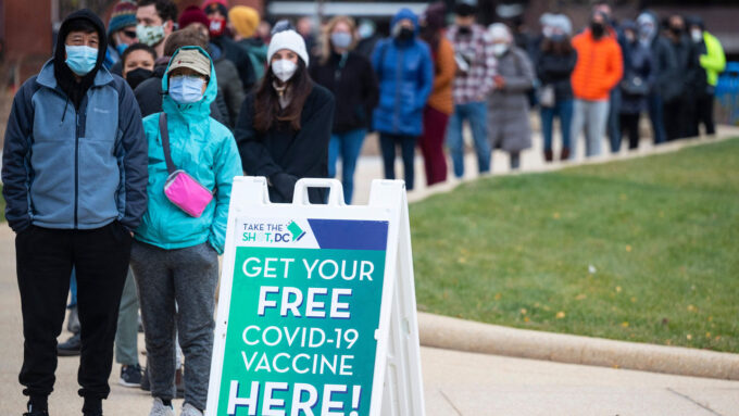 A line of people wearing masks wait in line behind a sign for free COVID-19 vaccines.