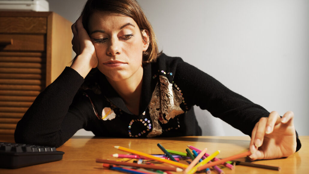 A woman representing procrastination as he slumps against a desk with her head in her hand and a pile of colored pencils in front of her.