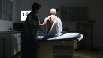 An elderly male patient sits on a medical exam bed with his back to the camera and a medical professional holding a stethoscope to the patient's back.