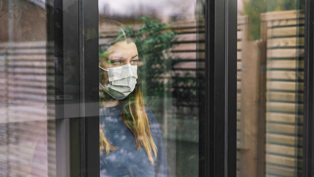 A woman wearing a face mask stands inside a home, looking out through a window