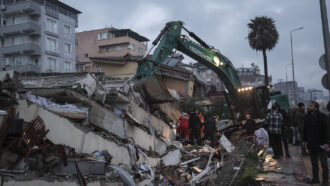 A photo of a green backhoe parked next to a pile of rubble while search and rescue workers stand nearby.