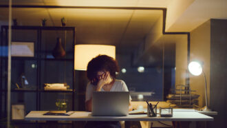 A photo of a woman sitting in front of a computer with her head in her hand, obscuring her face.