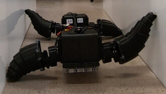 A photo of a new type of robot that has a black box for a body and four legs that are helping it move down the hall.
