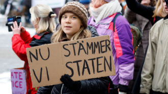 A photo of Greta Thunberg standing with a sign with a group of people standing behind her.