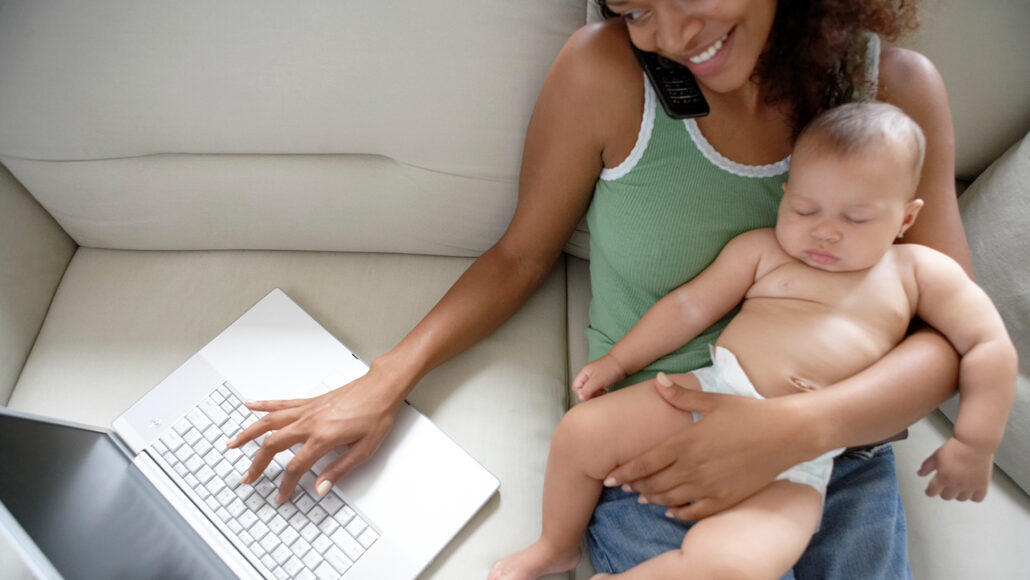 A photo of a woman sitting on a couch and holding a baby while she talks on the phone and types one handed on a laptop.
