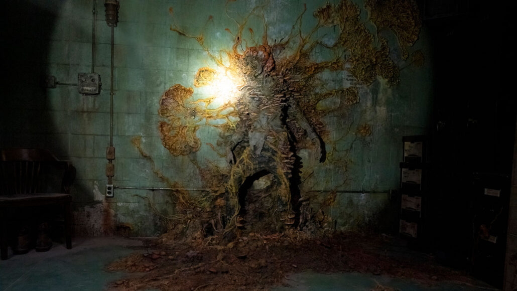 A still image from the television show The Last of Us showing a human body that has been completely covered by an orange fungus attached to a gray wall.