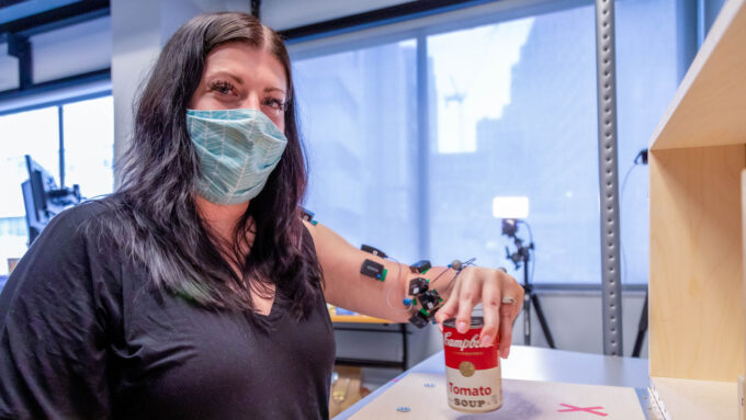 In a photo, stroke patient Heather Rendulic, who has dark hair and is wearing a dark shirt and a hospital mask, holds a Campbell's soup can. Devices used to monitor her implant are visible on her arm.