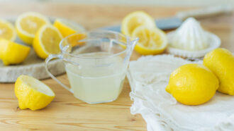 A photo of several lemons, some cut in half, and a small pitcher of lemon juice sitting on a wood counter top with a juicer in the background.