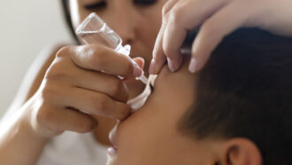 A photo of a boy with an eye drop bottle being held above his eye by his mom in the background.
