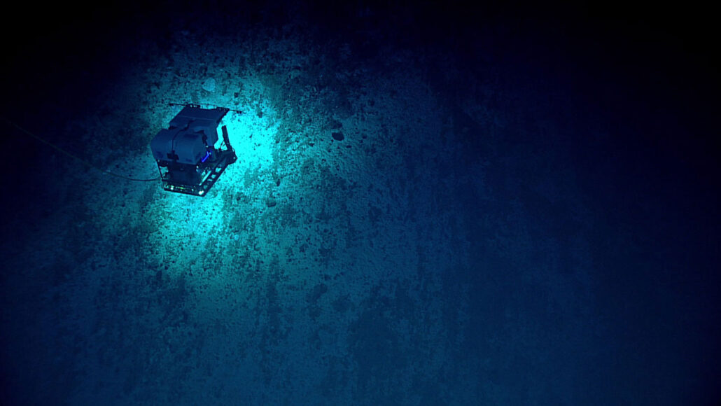 A photo of a remotely operated vehicle exploring the Mariana Trench. It appears as a cube hovering over a lighter blue patch surrounded by darkness.