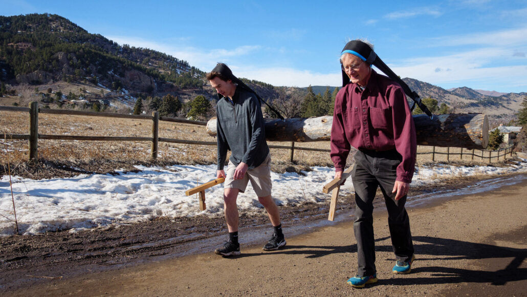 A photo of two men walking side by side on a road. Both men have a piece of fabric across their foreheads that attaches to a large log on their backs.