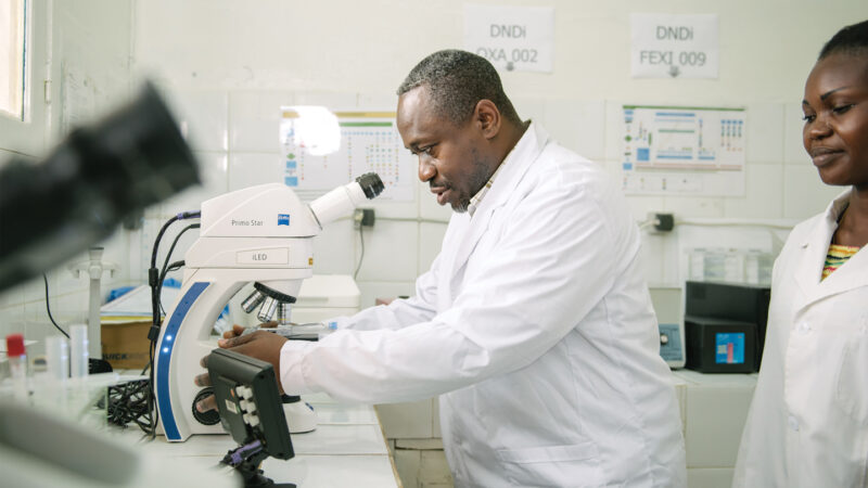 photo of Wilfried Mutombo Kalonji looking at a slide under a microscope as another scientist stands in the background