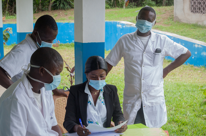 photo of nurse Béatrice Kasita holding some papers seated at a table outside a clinic surrounded by other healthcare workers