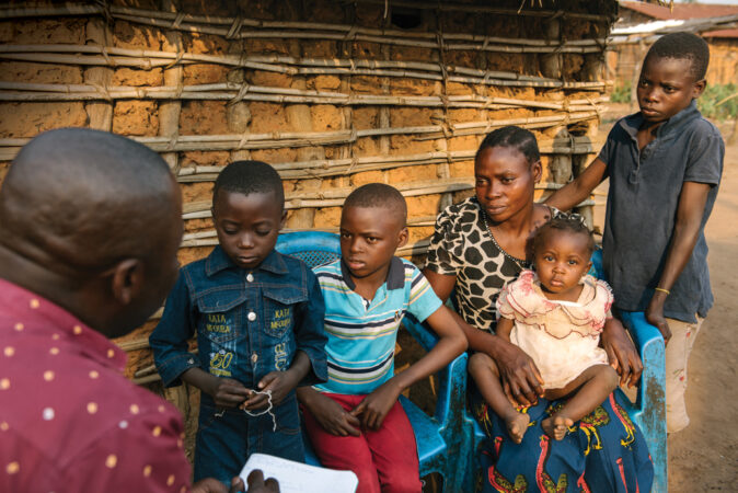 photo in which Felix Akwaso Massa appears in the foreground holding a notebook with his back to the camera as he speaks to Charnelle Mafuta and her four children who are seated in front of a building