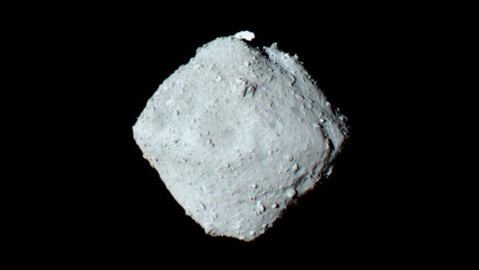 A photo take of the asteroid Ryugu. A light gray circular shape in the center on a deep black background.
