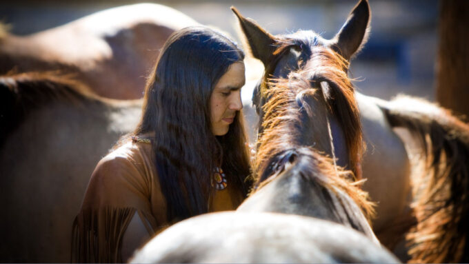 An image of a Native American man standing next to a brown horse while other brown horses mill in the background.