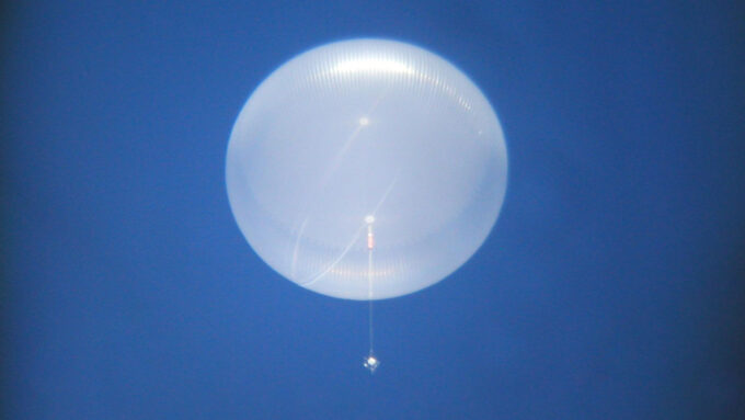 A photo of white opaque balloon with a wire and small contraption just below rising into a blue sky.