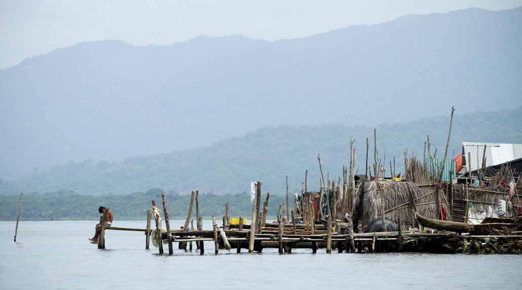 A far away photo of a person sitting on the end of a dock with a clutter of wooden docks and poles behind them.
