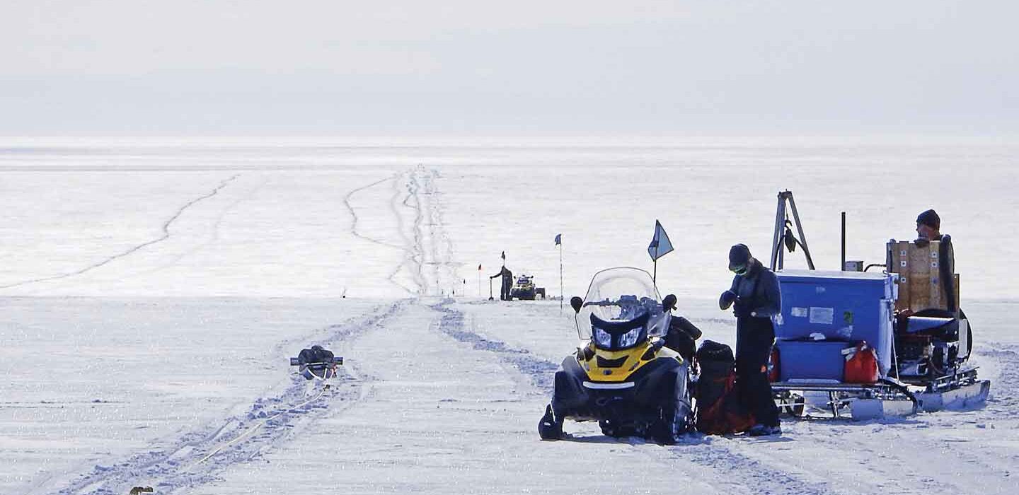 A photo of a person standing next to a snow mobile and a stack of supplies with tracks in the snow disappearing into the distance.