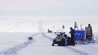 A photo of a person standing next to a snow mobile and a stack of supplies with tracks in the snow disappearing into the distance.