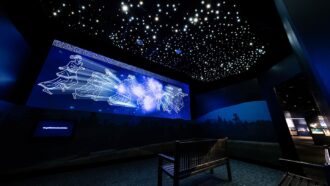 A photo of a dark room with a projector screen showing a film about the origin of a star cluster. The outlines of people and stars are on the screen frozen while stars light up the ceiling.
