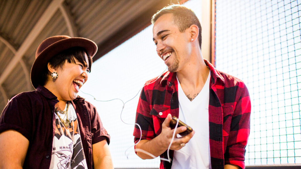 A photo of a man and a woman sharing a pair of headphones while listening to music on a handheld device.