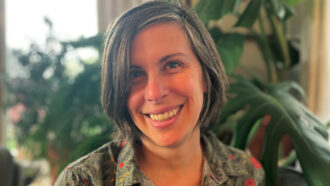 A headshot of Marjorie Weber smiling in front of large plants