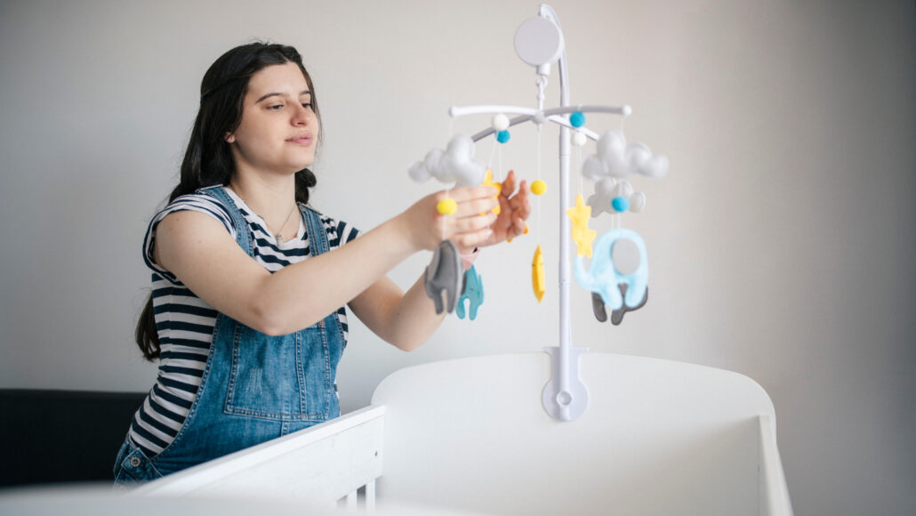 A photo of a dark haired woman adjusting a mobile over a baby's crib.