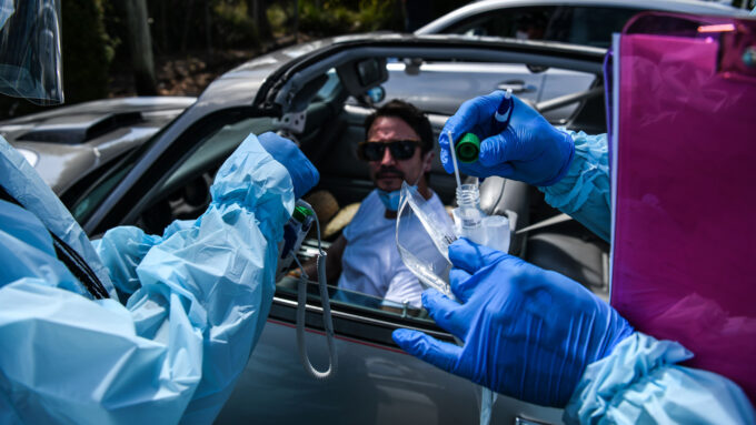 People wearing gloves and full-body personal protective equipment prepare a COVID-19 test for a person sitting in a car wearing sunglasses