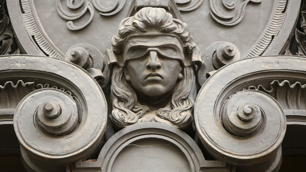 A close up photo of detail on a building showing the sculpture of a head with a blindfold covering the person's eyes.