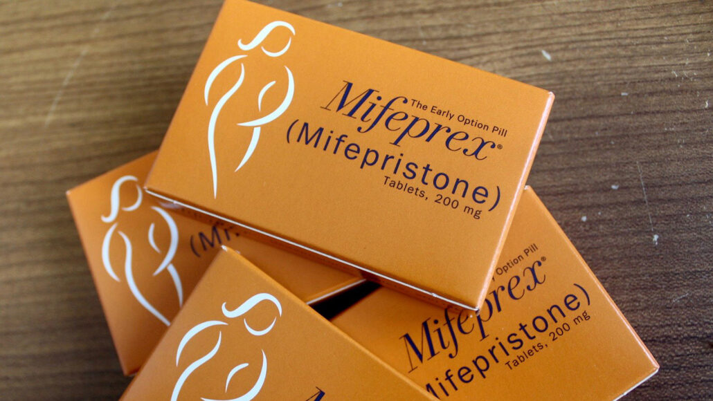 A photo of four orange Mifeprex pill boxes in a pile on a wooden background.