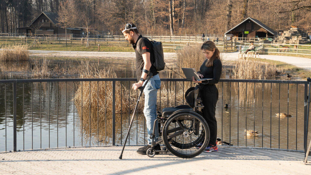 A photo of a man standing up from his wheelchair in front of a pond while a woman holds a laptop behind the wheelchair.